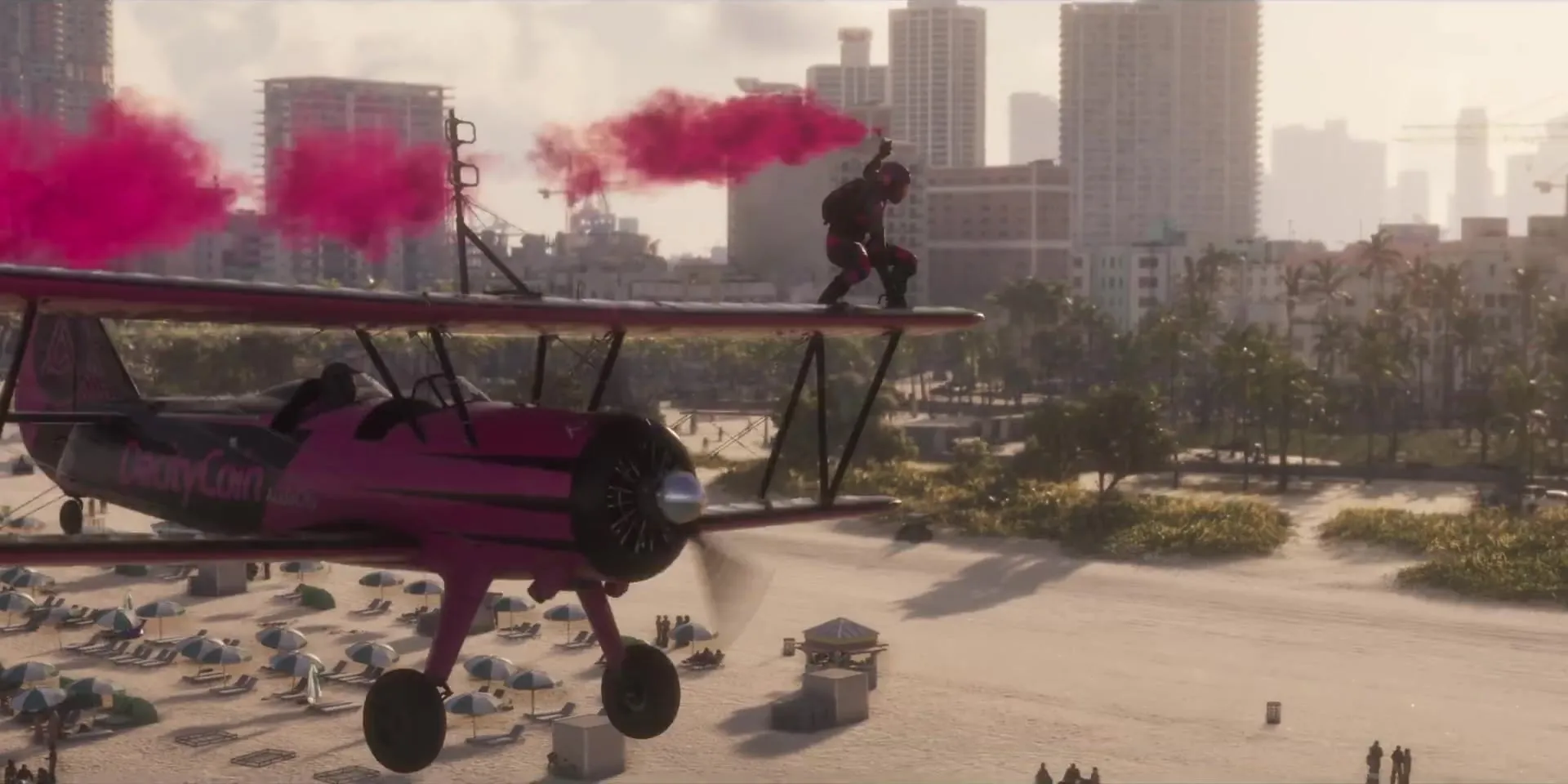 A pink biplane trailing pink smoke over a crowded beach, with a person standing on the wing