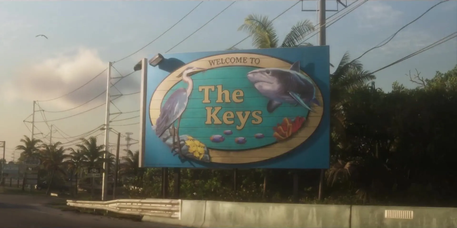 The Welcome to The Keys sign painted with a heron and a shark, along a palm-lined road