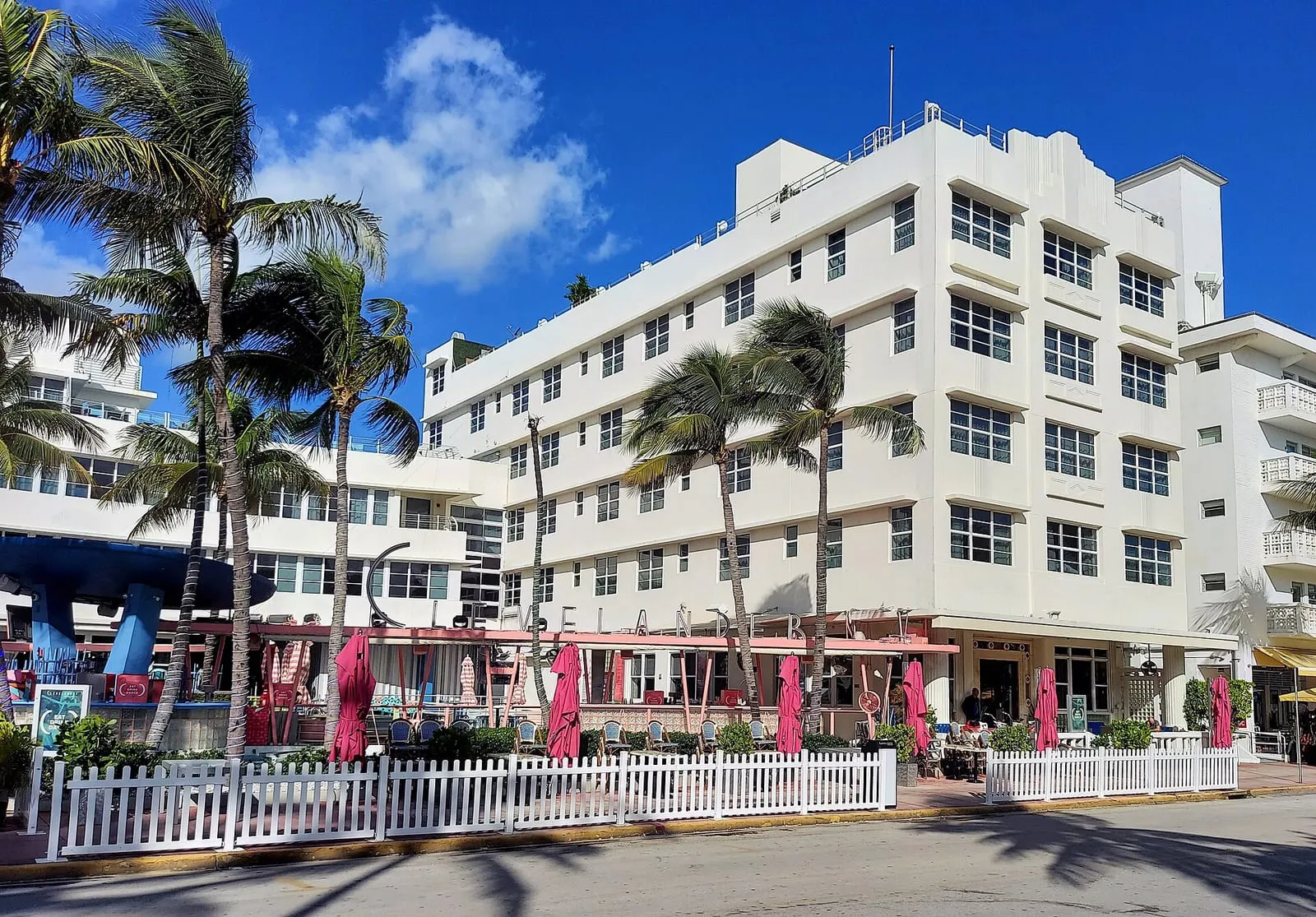 Il Clevelander Hotel di Miami Beach di giorno: palazzo bianco Art Deco su Ocean Drive con le palme, gli ombrelloni rosa del bar e la staccionata bianca sul marciapiede