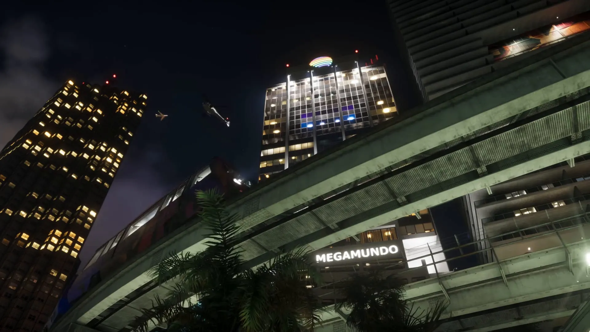 An Extended Look frame: the illuminated Megamundo sign at the base of a Vice City skyscraper at night, with a train passing on an elevated line and the rainbow-topped tower in the background